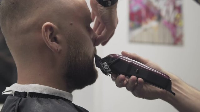 Close up of male barber trimming beard with shaver. Professional shaving bearded man in slow motion. Barber shaving beard with electric razor and comb in male salon.