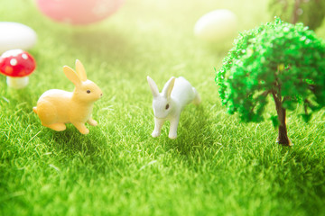 Cute Little Easter Bunnys in the Meadow. Spring Flowers and Green Grass. Sunbeams. Yellow easter egg spring meadow with two white rabbit and easter eggs. Fairy tale