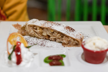 A piece of apple pie with ice cream and sauce on a white plate in a cozy cafe, close up
