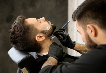 Professional hairdresser shaving client with straight razor in barbershop
