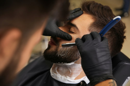 Professional hairdresser shaving client with straight razor in barbershop
