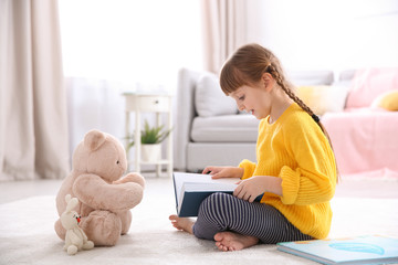 Cute little girl with teddy bear reading book on floor at home