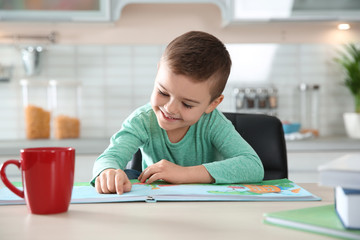 Cute little boy reading book at table in kitchen