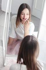 Pretty tween girl rouge lips holding red lipstick looking at big mirror sitting on floor in her room.