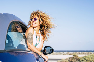 Beautiful cheerful happy caucasian woman outside the car with wind in curly long blonde hair - attractive people enjoying the freedom in outdoor coastline ocean vacation place