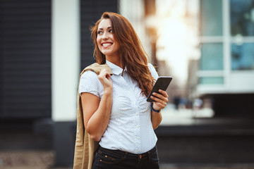 Beautiful young smiling woman checking social media on her smartphone against urban city background.