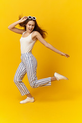 Fototapeta na wymiar Vertical image of trendy looking gorgeous emotional teenage girl wearing stylish summer clothes and sunglasses jumping or dancing against yellow studio wall background with copy space for your text