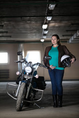 Portrait of Caucasian pregnant woman biker standing next to motorbike with white helmet in hand, underground parking