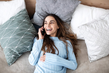 Beautiful smiling woman talking on cellphone while lying in cozy bed at home.
