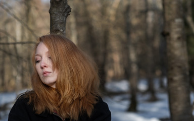 Young sexy woman with red hair and head down, with black clothes, is standing sad, depressed, desperate in the forest in winter.