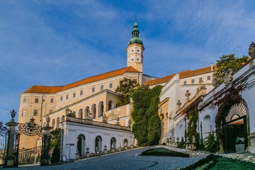 Mikulov, Czech Republic / South Moravia - October 15 2016: Mikulov castle with tower and yellow and white facade, white walls with statues, cobble stone pavement, sunny day, blue sky, green vegetation