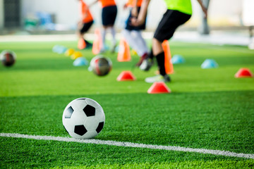 Obraz na płótnie Canvas football on green artificial turf with blurry soccer team training, blurry kid soccer player jogging between marker cones and control ball with soccer equipment in soccer academy.