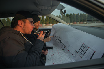 Two robbers sitting inside a car with guns looking at the blueprint of the building they are about...
