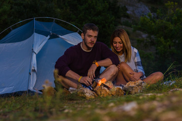 Young couple drinking coffee, enjoying the mountain view and warming up on the bonfire in the dusk