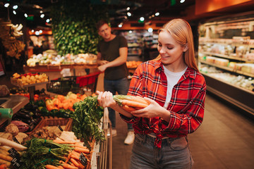 Young couple in grocery store. Woman stand in front and hold carrot in hands. She look at it and...