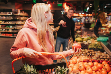 Young couple in grocery store. Woman hold pomegranat in hand and look at guy. He has grapes in hands. Smiling to each other.