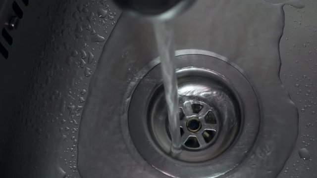 Top down view of running tap in stainless steel sink with water running down plug hole. Slow pan forward.