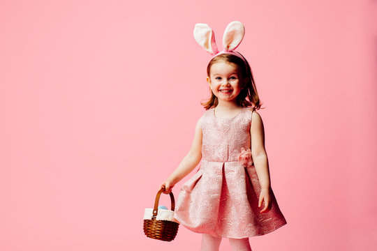 Portrait of a smiling little girl with easter eggs basket and bunny ears, on pink