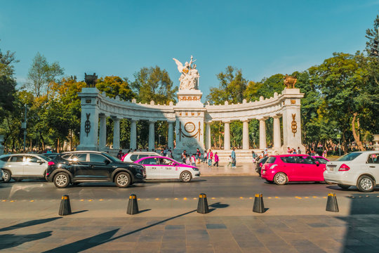  Fullshot view of a famous monument called "Hemiciclo a Juarez" located in Reforma, Mexico, surrounded by trees, cars and people walking on blue sky.