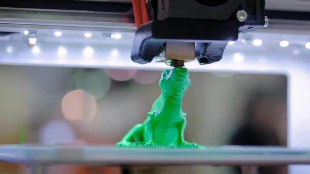 Print head of 3D printer machine printing plastic model of green toy lizard at modern scifi technology exhibition. 3D printing, additive technologies, 4.0 industrial revolution and futuristic concept