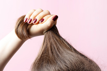 Female hands with red manicure holding hair, pink background, hair care concept
