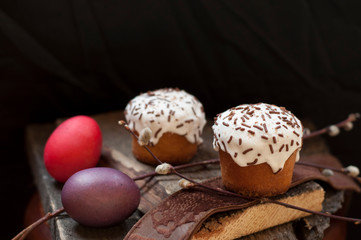 A  two small Easter cake and dyed  eggs, and a willow twig on a dark wooden background. Close up. Selective focus.