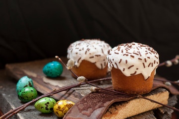 A  two small Easter cake and dyed quail eggs, and a willow twig on a dark wooden background. Close up. Selective focus.