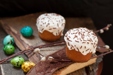 A  two small Easter cake and dyed quail eggs, and a willow twig on a dark wooden background. Close up. Selective focus.