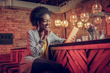 Beaming cheerful young-adult Afro-American woman enjoying hilarious video on tablet