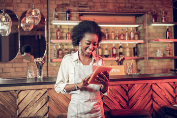 Smiling cheerful Afro-American bar waitress adding order information to tablet