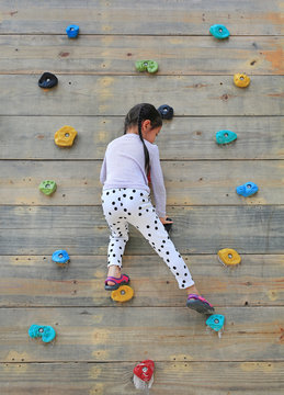 Little Child Girl Trying On Free Climbing On The Playground Wooden Wall Outdoors.