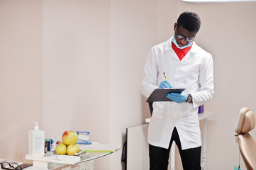 African american male doctor in glasses and mask posed in clinic with black clipboard at hand and writing something.