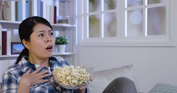 Surprised woman eating junk food on sofa at home watching horror movie. young asian lady spill popcorn while scared by thriller film. Terrified girl bowl of popcorn sit on couch hand touch heart.