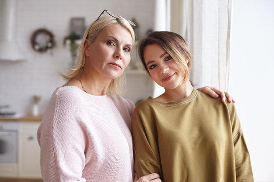 Picture of attractie blonde female in her forties embracing beautiful cute teenage daughter, being proud of her young child, posing in stylish cozy kitchen interior, looking at camera and smiling
