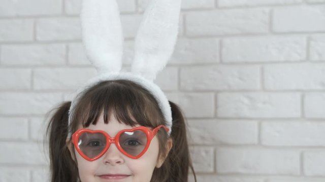 A child in the ears of a hare shows tongue. A cheerful girl in rabbit ears and sunglasses shows her tongue.