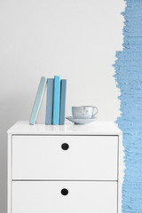 Books and cup with coffee on chest of drawers near wall
