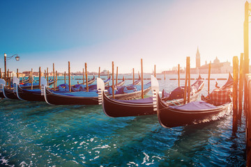 Gondolas stand in row blue-green water of Venetian canal, overexposure and glare on right, clear...