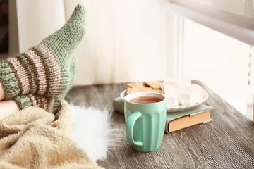 Foto op Plexiglas Cup of hot tea with legs of young woman on wooden table near window © Pixel-Shot