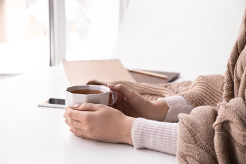 Young woman with cup of hot tea sitting at table near window at home