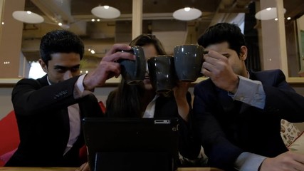 Handheld shot of coworkers colleagues huddled together at cafe table as they stare at laptop screen coming up with solution they cheers with their mugs together to celebrate and congratulate