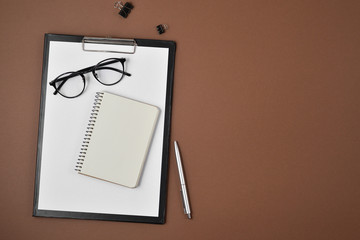 Mock up workspace with blank clip board, office supplies, pen, notepad and eyeglasses on brown...