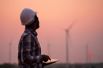 African engineer wearing white hard hat standing with digital tablet against wind turbine on sunny...