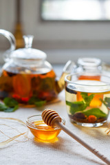 Tea with lemon and mint in a glass transparent tea pot, cup and honey in a bowl and a jar. Fresh mint and honey stick as decor. Healthy and delicious hot drink