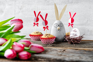 Lächelnder Dekohase zwischen Muffins und Tulpen wünscht Frohe Ostern