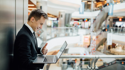 Cheerful young  economist  holding laptop and showing thumbs up, happy, dressed in black suit,...