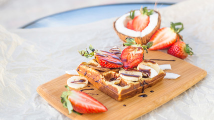 Belgian waffle with fruit and chocolate sauce is beautifully served on a wooden board. Next to strawberries and coconut
