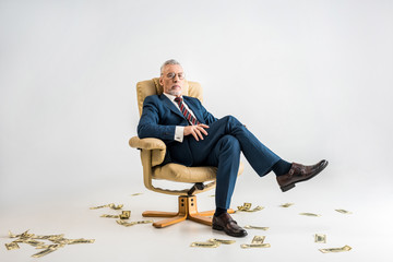 confident mature businessman sitting in armchair near dollar banknotes on grey