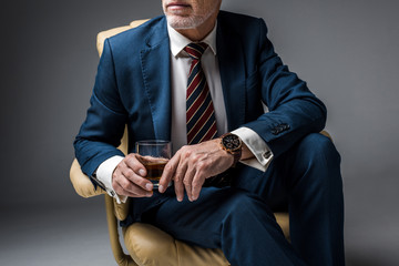 cropped view of mature businessman in suit sitting in armchair and holding glass of whiskey on grey
