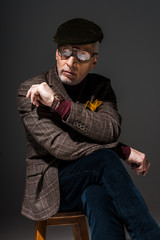 Obraz premium pensive mature man in glasses and cap sitting on chair and thinking on grey