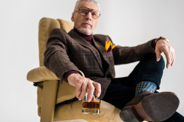 Obraz premium selective focus of mature man sitting in armchair with crossed legs and holding glass of whiskey isolated on grey